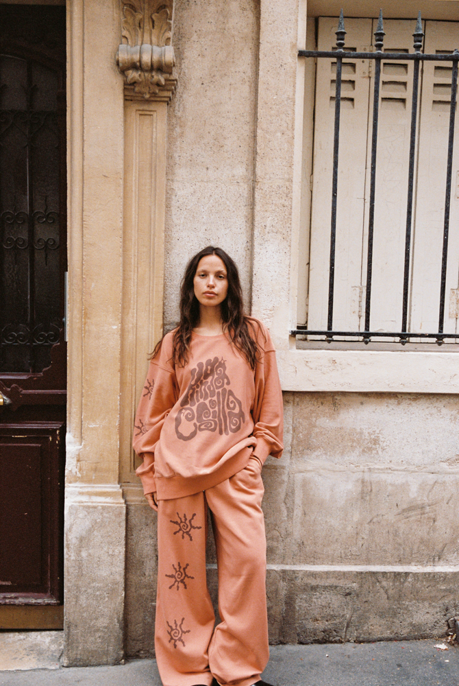 Model leaning against a stone wall in Paris wearing Kinga Csilla Abracadabra Hades Pant with matching Abracadabra OG Jumper in burnt orange with sun motif