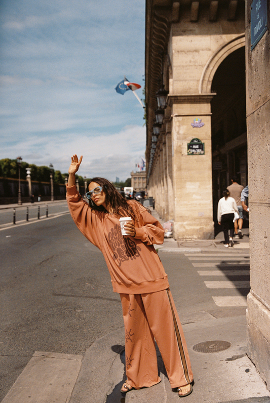 Model wearing Kinga Csilla Abracadabra Hades Pant with matching Abracadabra  OG Jumper in burnt orange, standing on Rue de Rivoli in Paris, waving with coffee in hand
