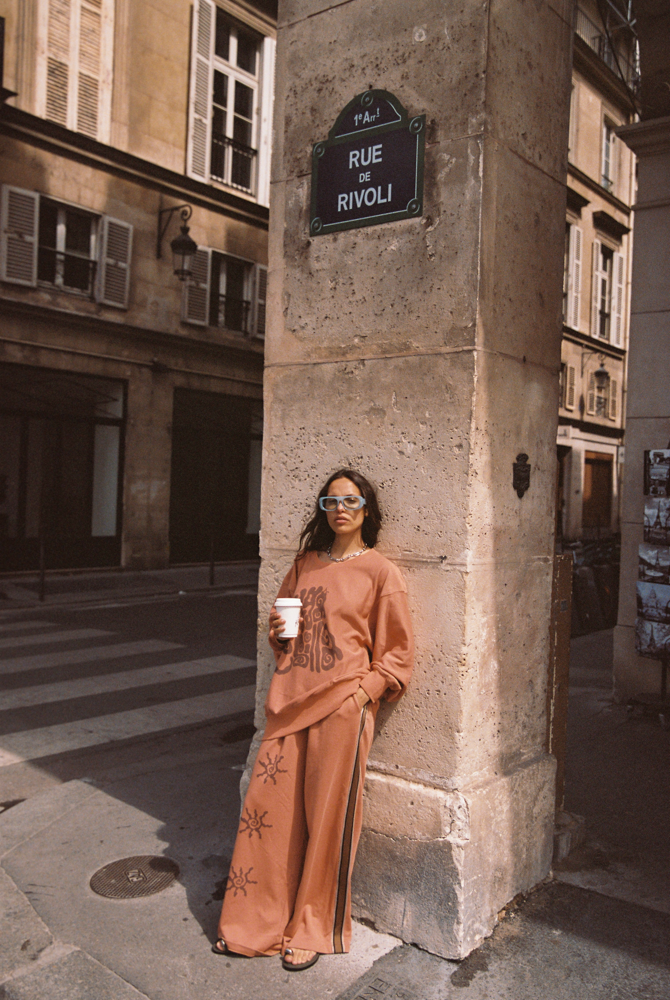 Model leaning against stone pillar under Rue de Rivoli street sign in Paris wearing Kinga Csilla Abracadabra Hades Pant and Abracadabra OG Jumper in burnt orange with sun print