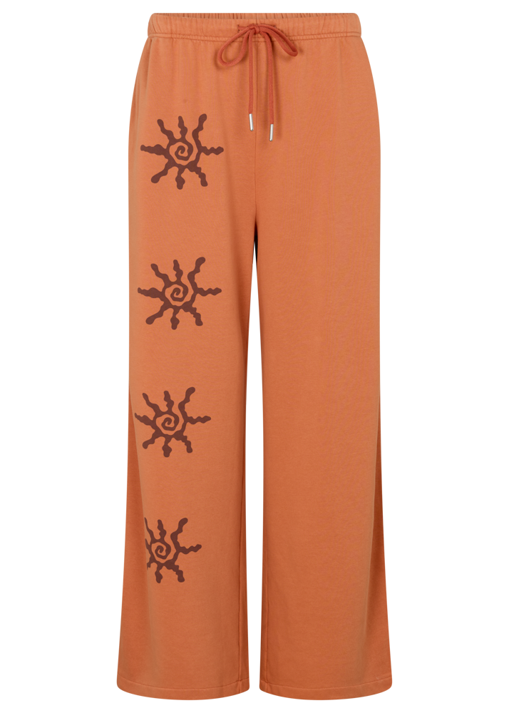 Flat lay of Kinga Csilla Abracadabra Hades Pant in bright orange with sun spiral graphic print on right leg, featuring elasticated waistband, drawstring, and flared wide-leg hem