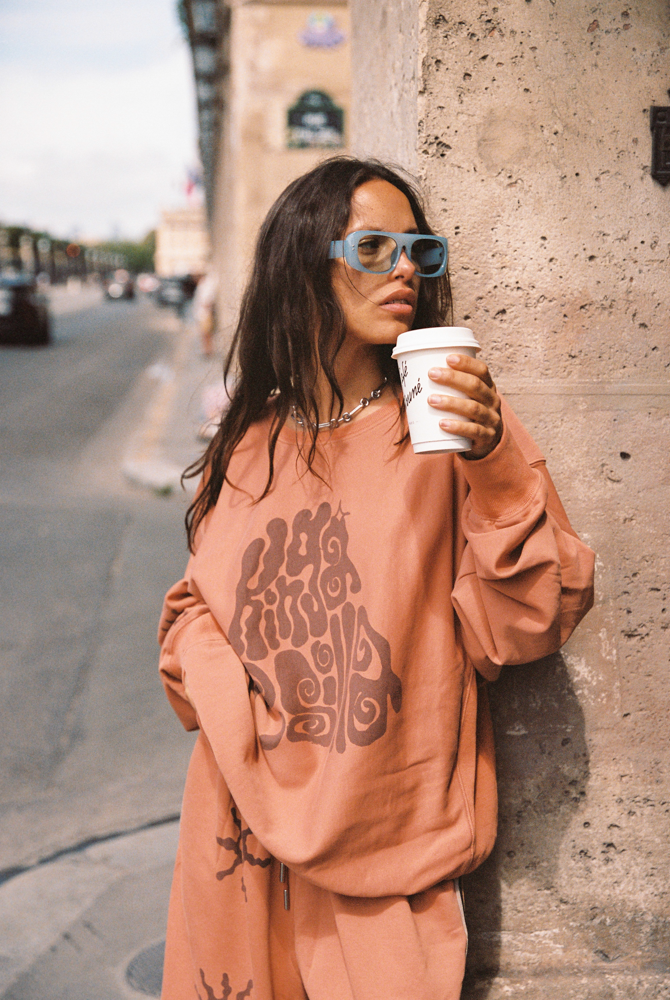 Model wearing Kinga Csilla Abracadabra OG Jumper in warm orange with oversized fit and graphic print, styled street-side with coffee cup and sunglasses