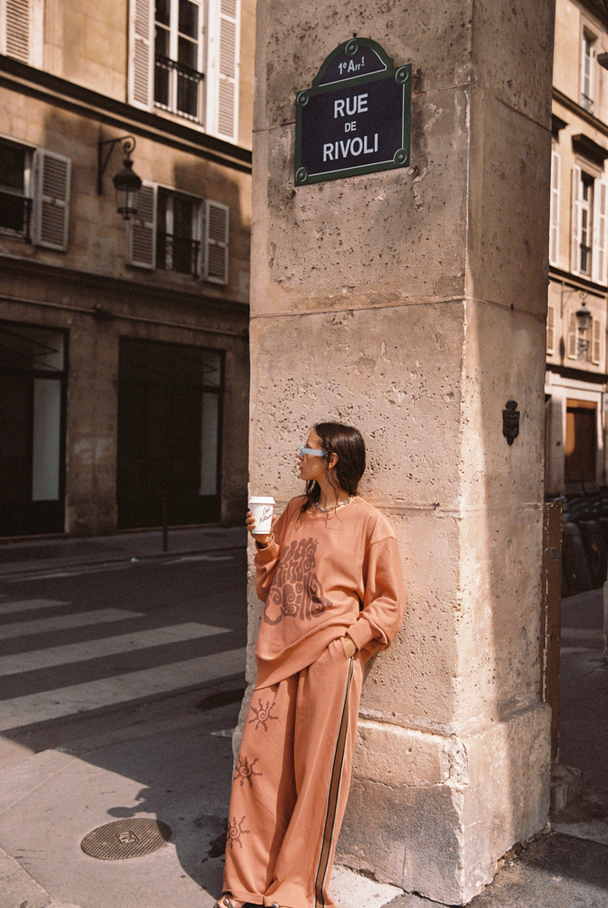Model wearing Kinga Csilla Abracadabra OG Jumper in terracotta orange with wide-leg Abracadabra  Hades Pants, styled on Rue de Rivoli in Paris with oversized streetwear look and coffee cup