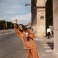 Model in Kinga Csilla Abracadabra OG Jumper and matching wide-leg Abracadabra Hades Pants in terracotta orange, posing playfully on Rue de Rivoli in Paris with coffee cup and sunglasses