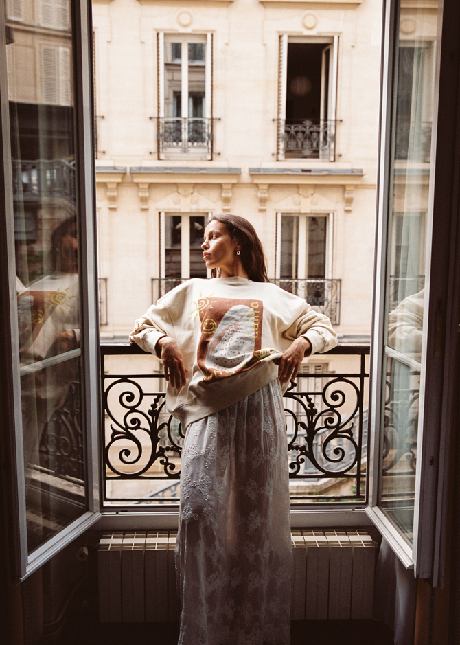 Woman standing on a Parisian balcony in the Kinga Csilla Shell OG Jumper cream oversized sweater, styled with a white lace maxi skirt for a relaxed bohemian outfit.