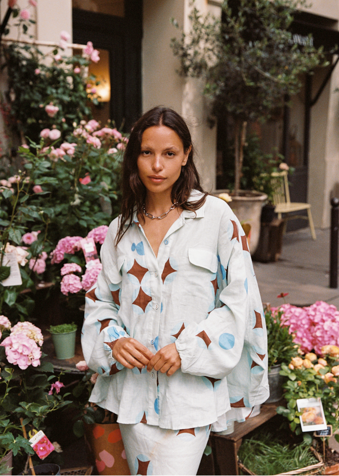Model wearing Kinga Csilla Aquarius Chalm Shirt in baby blue linen with celestial brown and blue print, styled with matching Aquarius Column Skirt among pink flowers