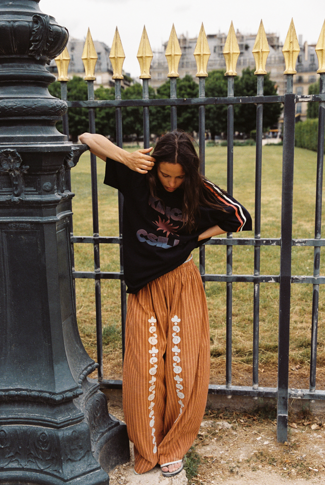 Model in Kinga Csilla Bello Swirl Jessi Pant with white embroidery, styled with oversized Kinga Csilla Sunset tee, leaning against Paris fence in casual street style setting