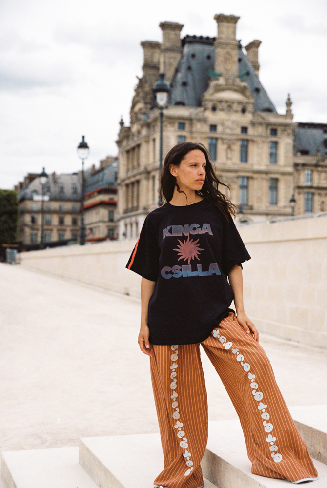 Model wearing Kinga Csilla Bello Swirl Jessi Pant in orange with white embroidery, styled with oversized Kinga Csilla Sunset tee in Paris street style setting