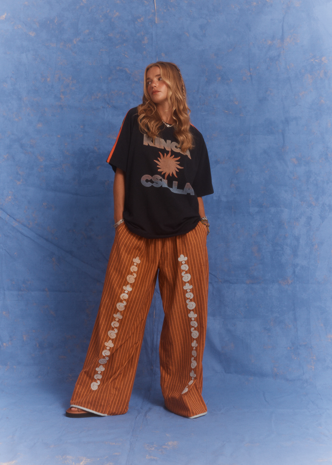 Model wearing Kinga Csilla oversized Sunset t-shirt in black with burnt orange stripe detail, styled with wide leg Bello Swirl Jessi Pant featuring white embroidery