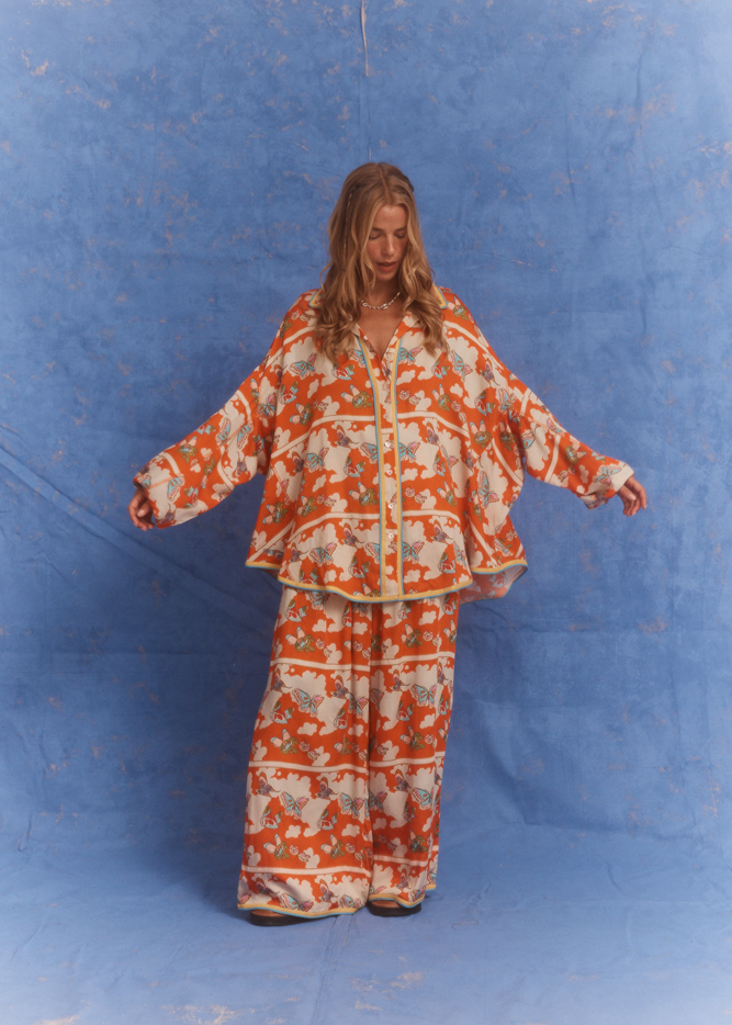 Model wearing Kinga Csilla Farfalla Chalm Shirt and Jessi Pant in oversized orange butterfly print co-ord set against blue backdrop