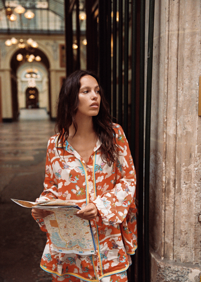 Model wearing the Kinga Csilla Farfalla Chalm Shirt in orange butterfly print, styled with matching Farfalla Jessi Pant, holding a map in a Paris arcade.