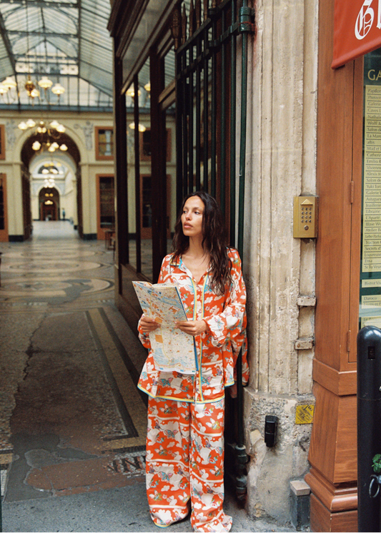 Model wearing Kinga Csilla Farfalla Jessi Pant with matching Farfalla Chalm Shirt—relaxed wide-leg set in orange butterfly print—holding a city map inside a Paris covered arcade.