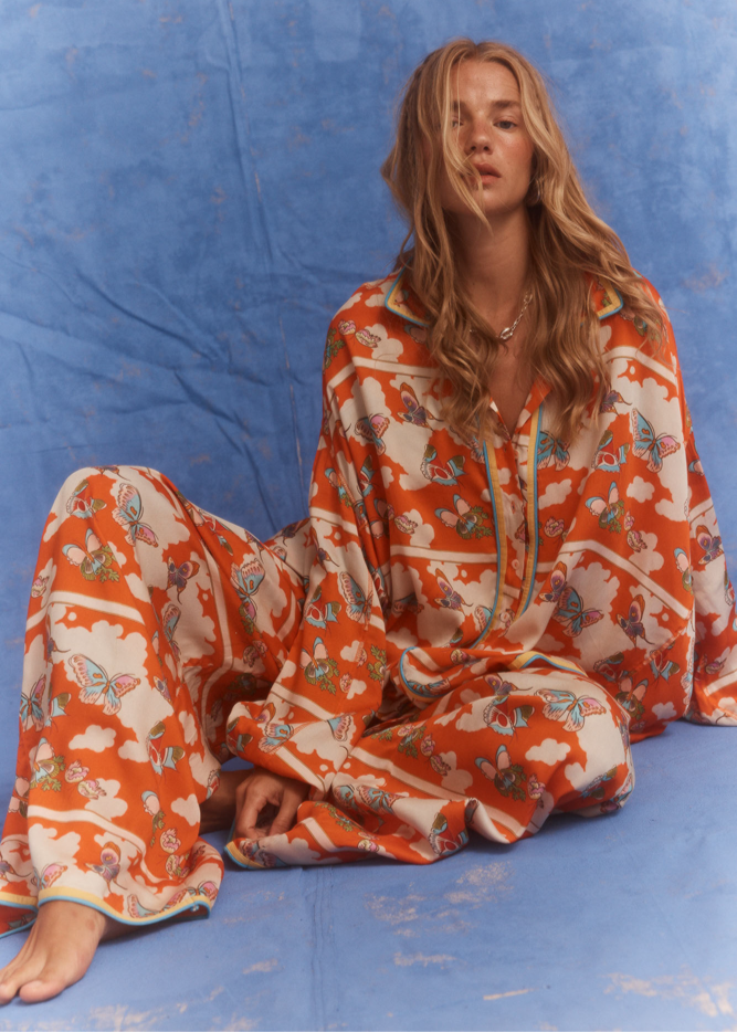 Kinga Csilla Farfalla Jessi Pant in orange butterfly print, relaxed wide-leg women’s pants styled with matching Farfalla Chalm Shirt, model seated against blue backdrop.