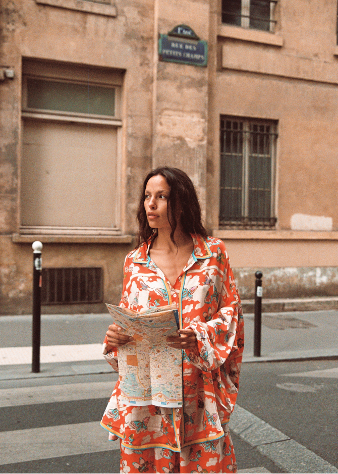 Model in Kinga Csilla Farfalla Jessi Pant with matching Farfalla Chalm Shirt, orange butterfly print set, holding a map while standing on a Paris street.