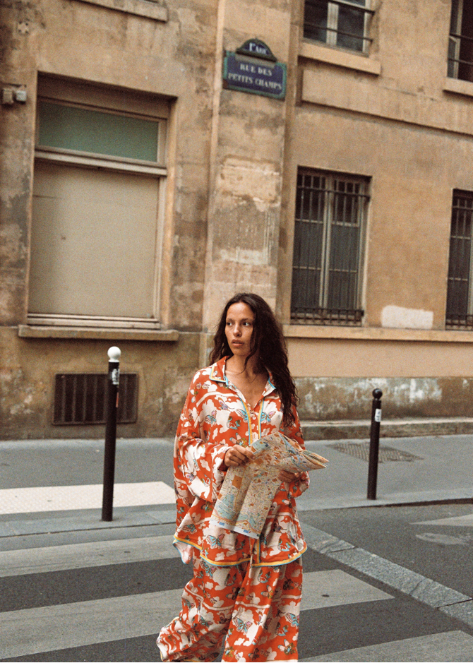 Model in Paris wearing Kinga Csilla Farfalla Jessi Pant and Farfalla Chalm Shirt in vibrant orange butterfly print, holding a city map while walking across a pedestrian crossing.