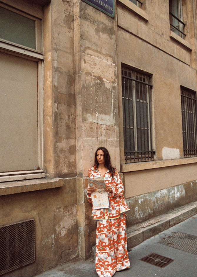 Model in Kinga Csilla Farfalla Jessi Pant and Farfalla Chalm Shirt, leaning against a Parisian stone wall while holding a city map, dressed in a bold orange butterfly print co-ord.