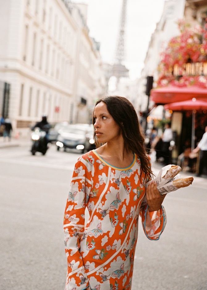 Model wearing Kinga Csilla Farfalla Olive Dress, a long-sleeve orange maxi with butterfly and floral print, holding baguettes on a Paris street with the Eiffel Tower blurred in the background.
