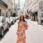 Full-length view of model in Kinga Csilla Farfalla Olive Dress, a long-sleeve orange maxi with butterfly and floral print, walking down a Paris street holding baguettes, Eiffel Tower in the distance.