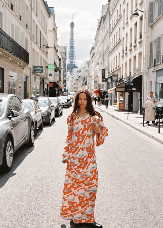 Full-length view of model in Kinga Csilla Farfalla Olive Dress, a long-sleeve orange maxi with butterfly and floral print, walking down a Paris street holding baguettes, Eiffel Tower in the distance.