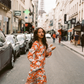 Model walking down a Paris street in the Kinga Csilla Farfalla Olive Dress, a long-sleeve orange maxi with butterfly and floral print, holding fresh baguettes with the Eiffel Tower in the background.