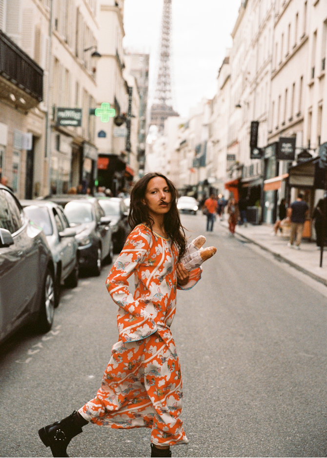 Model walking down a Paris street in the Kinga Csilla Farfalla Olive Dress, a long-sleeve orange maxi with butterfly and floral print, holding fresh baguettes with the Eiffel Tower in the background.