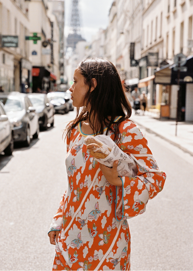 Model wearing the Kinga Csilla Farfalla Olive Dress, a flowing orange maxi with butterfly and floral motifs, carrying baguettes on a sunlit Paris street with the Eiffel Tower behind.
