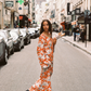 Model wearing the Kinga Csilla Farfalla Olive Dress, a flowing orange maxi with butterfly print, walking down a Paris street holding baguettes with the Eiffel Tower in the background.