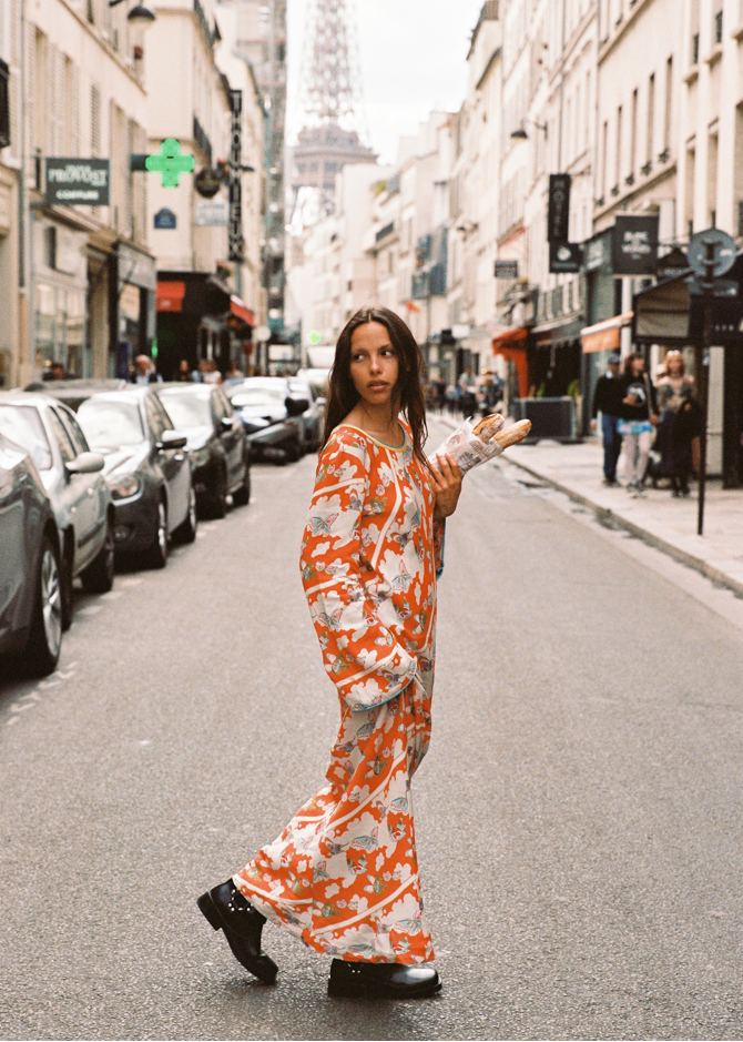 Model wearing the Kinga Csilla Farfalla Olive Dress, a flowing orange maxi with butterfly print, walking down a Paris street holding baguettes with the Eiffel Tower in the background.