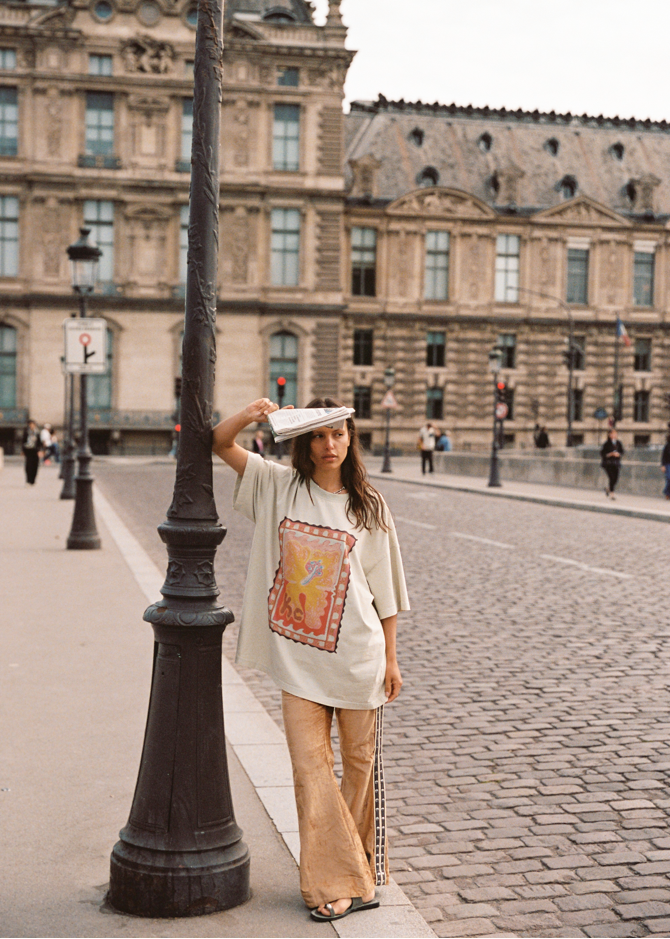 Model wearing Kinga Csilla Hibiscus Tee with bold hibiscus print, styled with TWILIGHT VELVET FLARE PANT, standing on Paris bridge with newspaper in hand