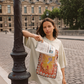 Model wearing oversized Kinga Csilla Hibiscus Tee with vintage stamp-style hibiscus print, holding a newspaper on Parisian street
