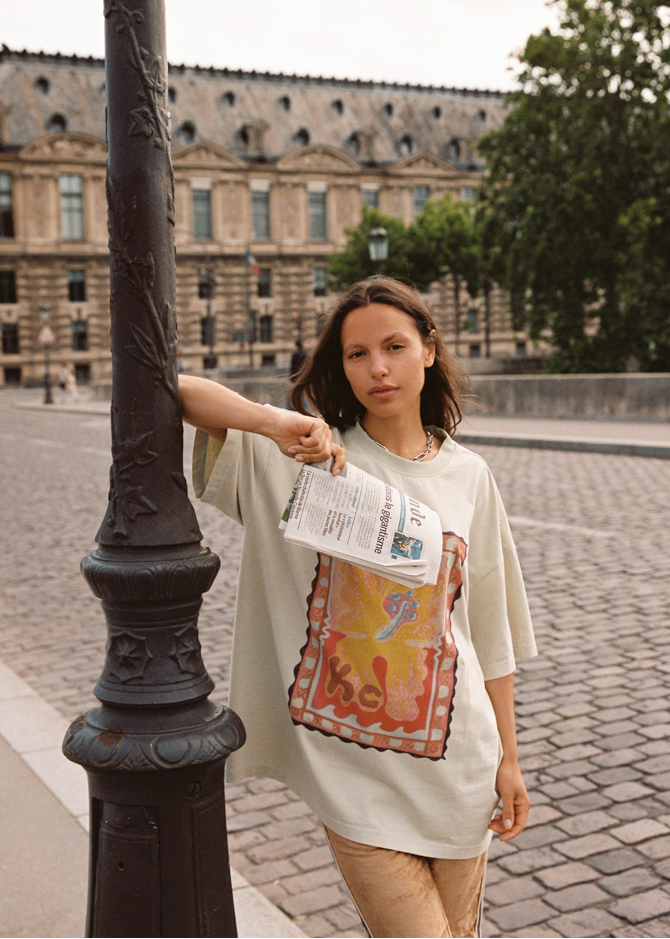Model wearing oversized Kinga Csilla Hibiscus Tee with vintage stamp-style hibiscus print, holding a newspaper on Parisian street