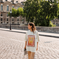 Model wearing Kinga Csilla HIBISCUS TEE, oversized off-white tee with vintage hibiscus print, styled with TWILIGHT VELVET FLARE PANT, holding newspaper while walking across cobblestone bridge in Paris.