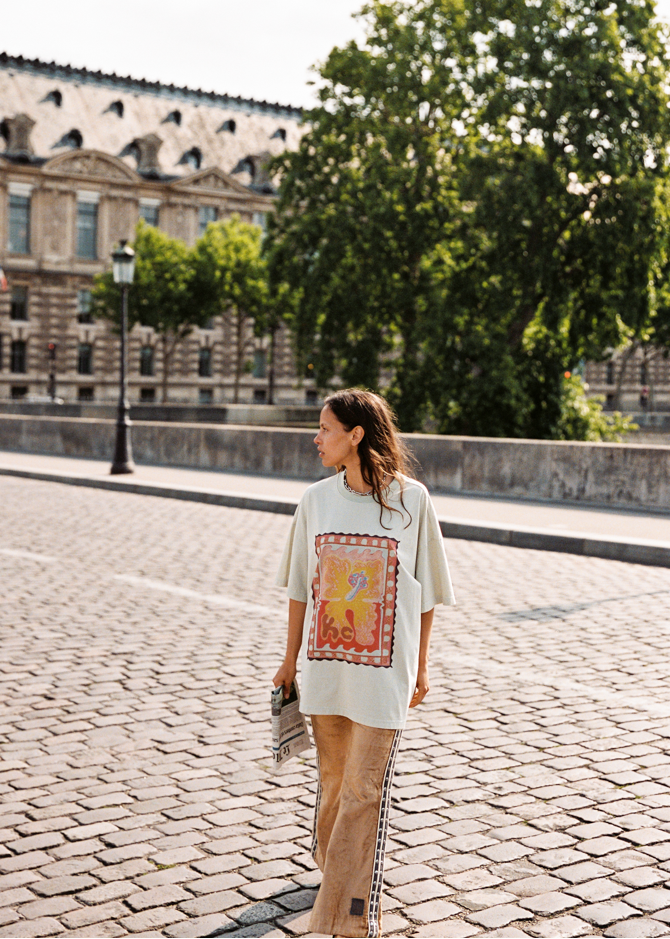 Model wearing Kinga Csilla HIBISCUS TEE, oversized off-white tee with vintage hibiscus print, styled with TWILIGHT VELVET FLARE PANT, holding newspaper while walking across cobblestone bridge in Paris.