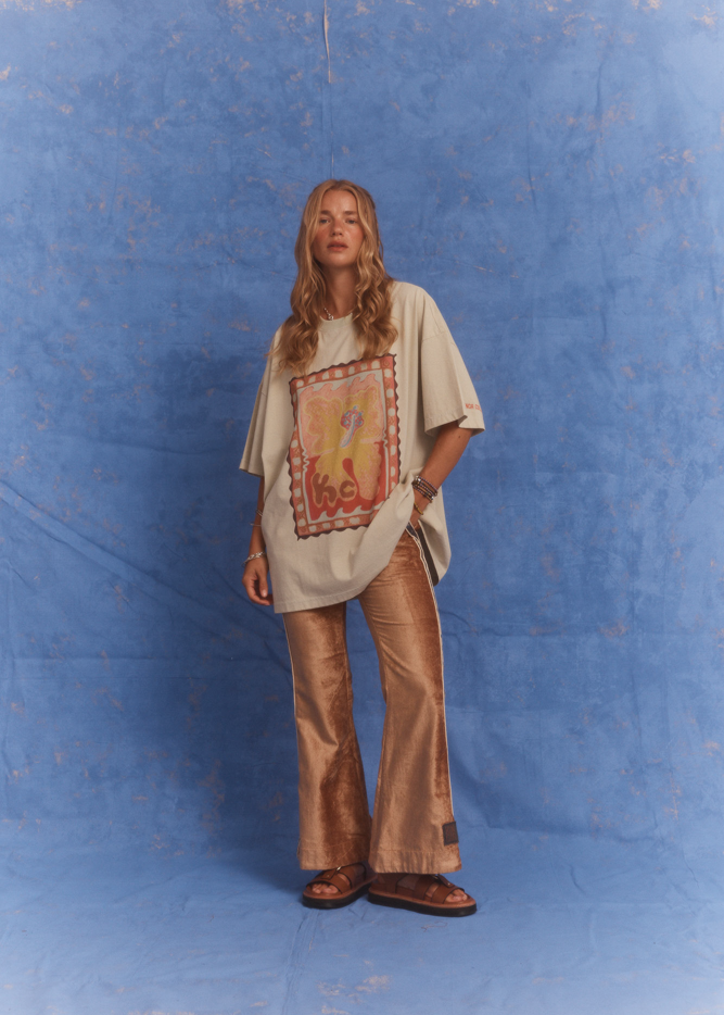 Model wears the Kinga Csilla Hibiscus Tee in studio against a blue backdrop, styled oversized with TWILIGHT VELVET FLARE PANT