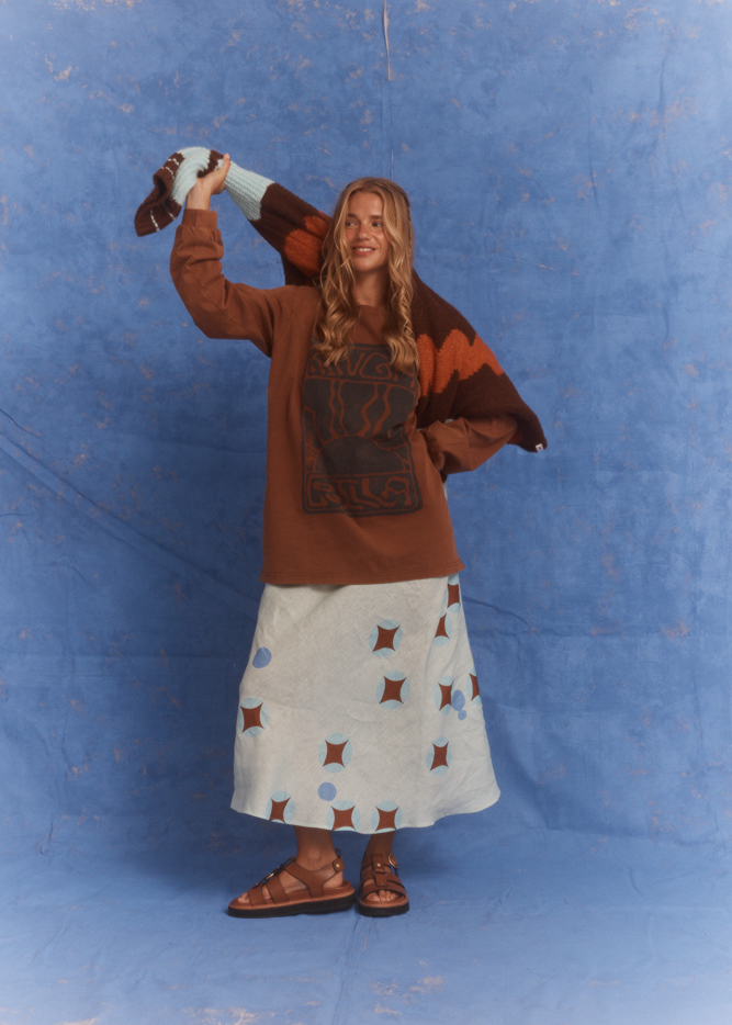 Model wearing Kinga Csilla Lino Roses Tee brown long sleeve styled with Aquarius Slip Dress, brown sandals and Surf Citta Cardigan draped over shoulders, styled in front of a blue studio backdrop