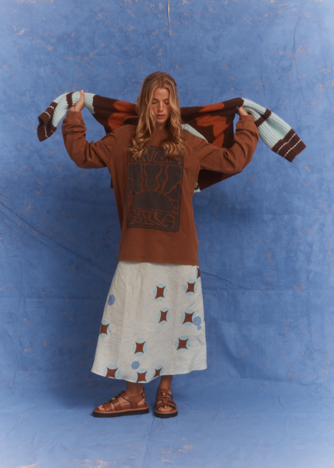 Model wearing Kinga Csilla Lino Roses Tee brown long sleeve styled with Aquarius Slip Dress and brown sandals, holding Surf itty Cardigan above shoulders, photographed in front of blue backdrop