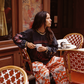 Model wearing the Kinga Csilla Love Potion Roses Tee in stonewashed black long sleeve, styled with Farfalla Pants, sitting at a Parisian café. Sustainable bohemian fashion look.