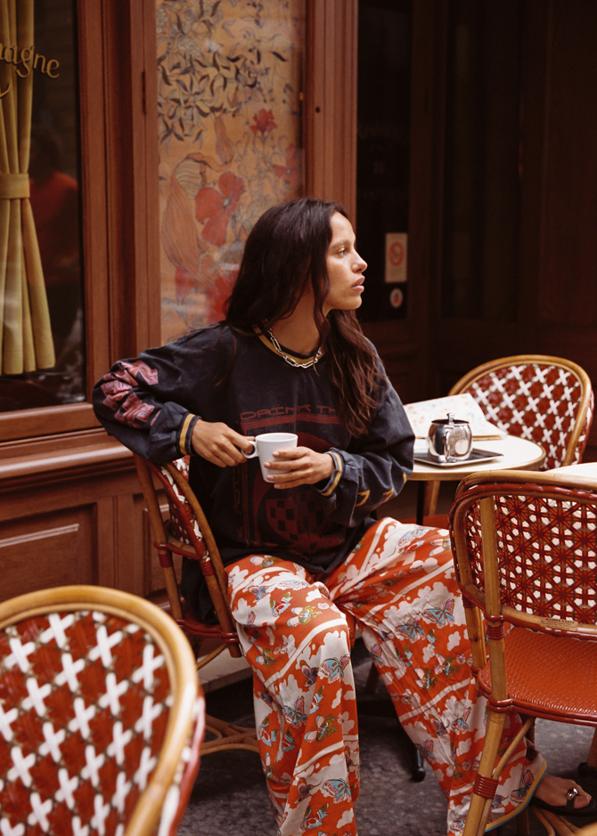 Model wearing the Kinga Csilla Love Potion Roses Tee in stonewashed black long sleeve, styled with Farfalla Pants, sitting at a Parisian café. Sustainable bohemian fashion look.