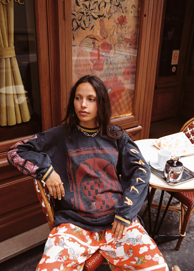 Model wearing Kinga Csilla Love Potion Roses Tee in stonewashed black with graphic rose print, styled with Farfalla Jessi Pant in orange butterfly cloud print, sitting at a Parisian café