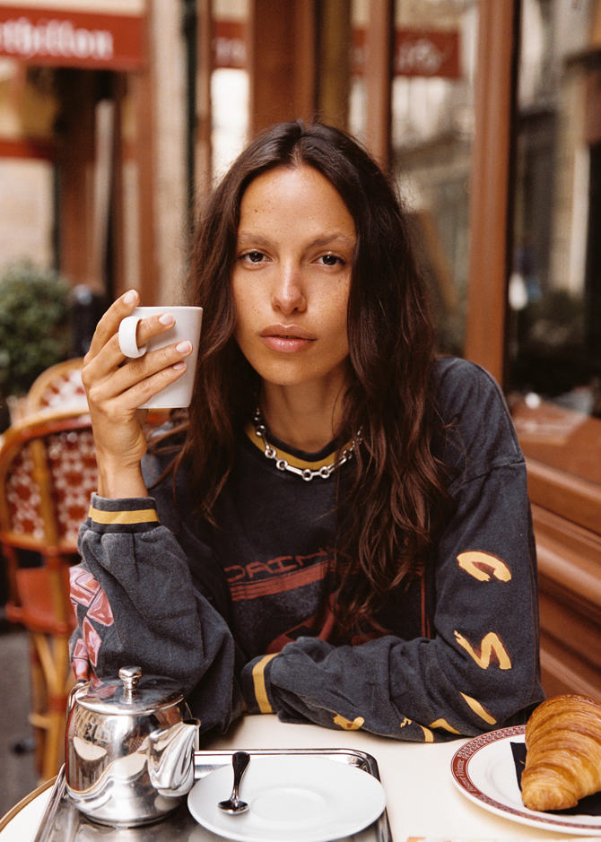 Close-up of model wearing the Kinga Csilla Love Potion Roses Tee in stonewashed black oversized long sleeve, holding a coffee at a Paris café. Sustainable bohemian street style.