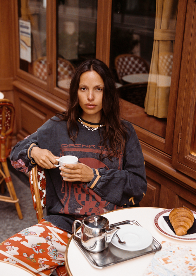 Model wearing the Kinga Csilla Love Potion Roses Tee in stonewashed black oversized long sleeve at a Paris café, styled with Farfalla print pants. Relaxed sustainable streetwear look.