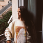 Model leaning on a balcony railing wearing the Kinga Csilla Shell OG Jumper in cream, oversized cotton jersey sweater with shell and sun doodles, styled in bohemian streetwear.