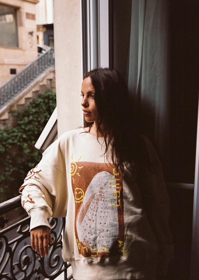 Model leaning on a balcony railing wearing the Kinga Csilla Shell OG Jumper in cream, oversized cotton jersey sweater with shell and sun doodles, styled in bohemian streetwear.