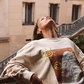 Model wearing the Kinga Csilla Shell OG Jumper, a cream oversized cotton sweater with hand-drawn shell and sun doodle graphic, styled casually outdoors on a Parisian-style balcony.