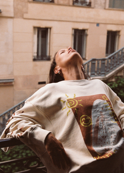 Model wearing the Kinga Csilla Shell OG Jumper, a cream oversized cotton sweater with hand-drawn shell and sun doodle graphic, styled casually outdoors on a Parisian-style balcony.