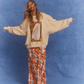Kinga Csilla Shell OG Jumper in cream, oversized fit with shell and sun doodle graphic, styled with vibrant orange floral Frafalla Olive Dress for a relaxed bohemian look against a blue backdrop.