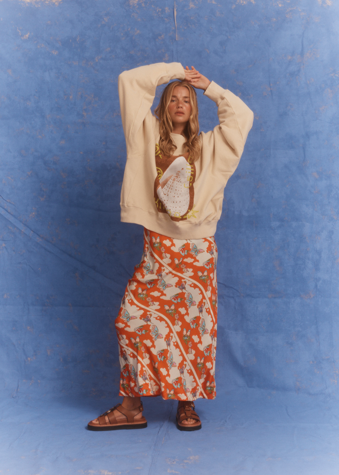 Model wearing the Kinga Csilla Shell OG Jumper, a cream oversized cotton sweater with hand-drawn shell and sun doodle graphic, styled with bold orange floral Farfalla Olive Dress and brown sandals. Relaxed bohemian streetwear look against a textured blue backdrop.