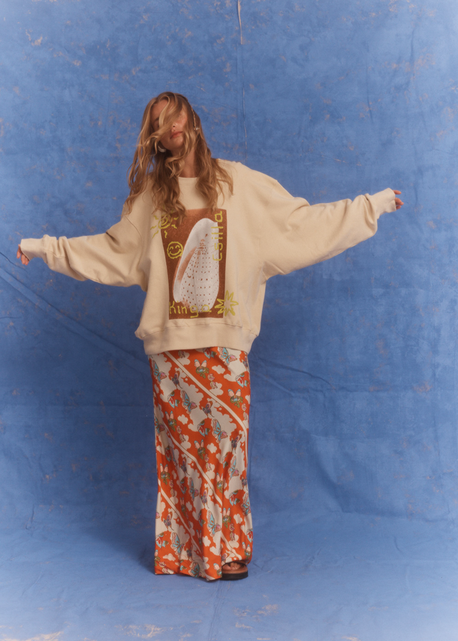 Kinga Csilla Shell OG Jumper in cream, oversized fit with shell and sun doodle graphic, styled with vibrant orange floral Frafalla Olive Dress for a relaxed bohemian look against a blue backdrop.