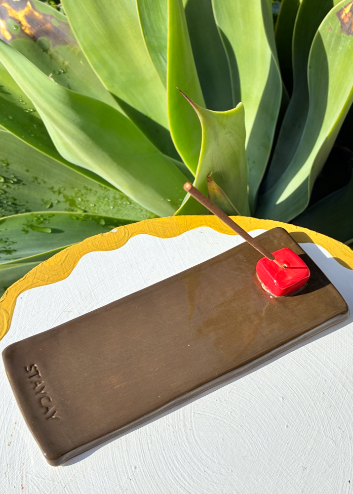 Handcrafted Kinga Csilla x Staycay ceramic incense holder in earthy brown with a glossy red cube incense rest. Unique bohemian artisan incense tray for home décor, meditation, and aromatherapy. Styled outdoors on a white scalloped table with lush green succulent backdrop.