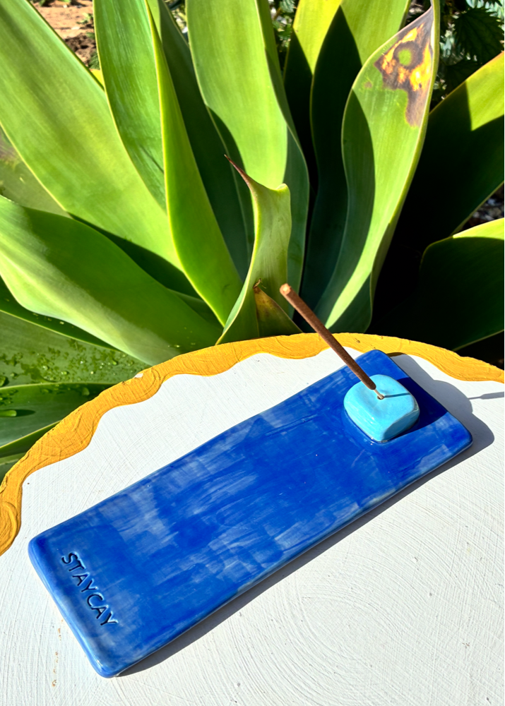 Deep blue Kinga Csilla x Staycay ceramic incense holder with a glossy sky blue cube incense rest. Handmade artisan incense tray for meditation, rituals, and home styling. Photographed outdoors on a scalloped white and yellow table with lush green plant background.
