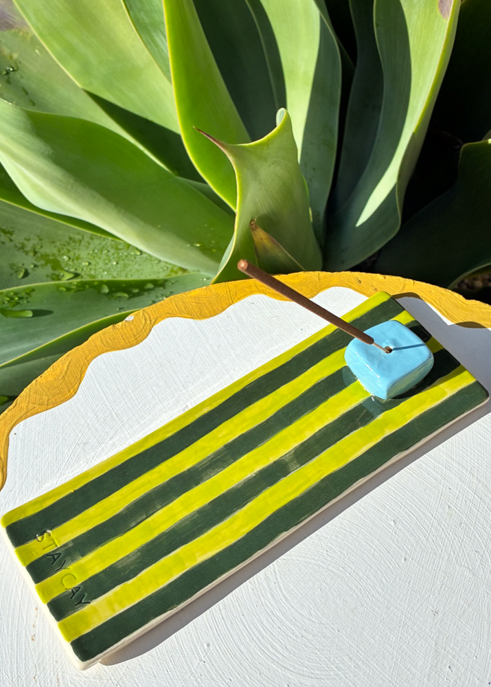 Handmade Kinga Csilla x Staycay ceramic incense holder with bold green and yellow stripes, finished with a glossy sky blue cube incense rest. Outdoor photo with incense stick on white scalloped table against green succulent leaves. Unique artisan bohemian home décor accessory for aromatherapy.
